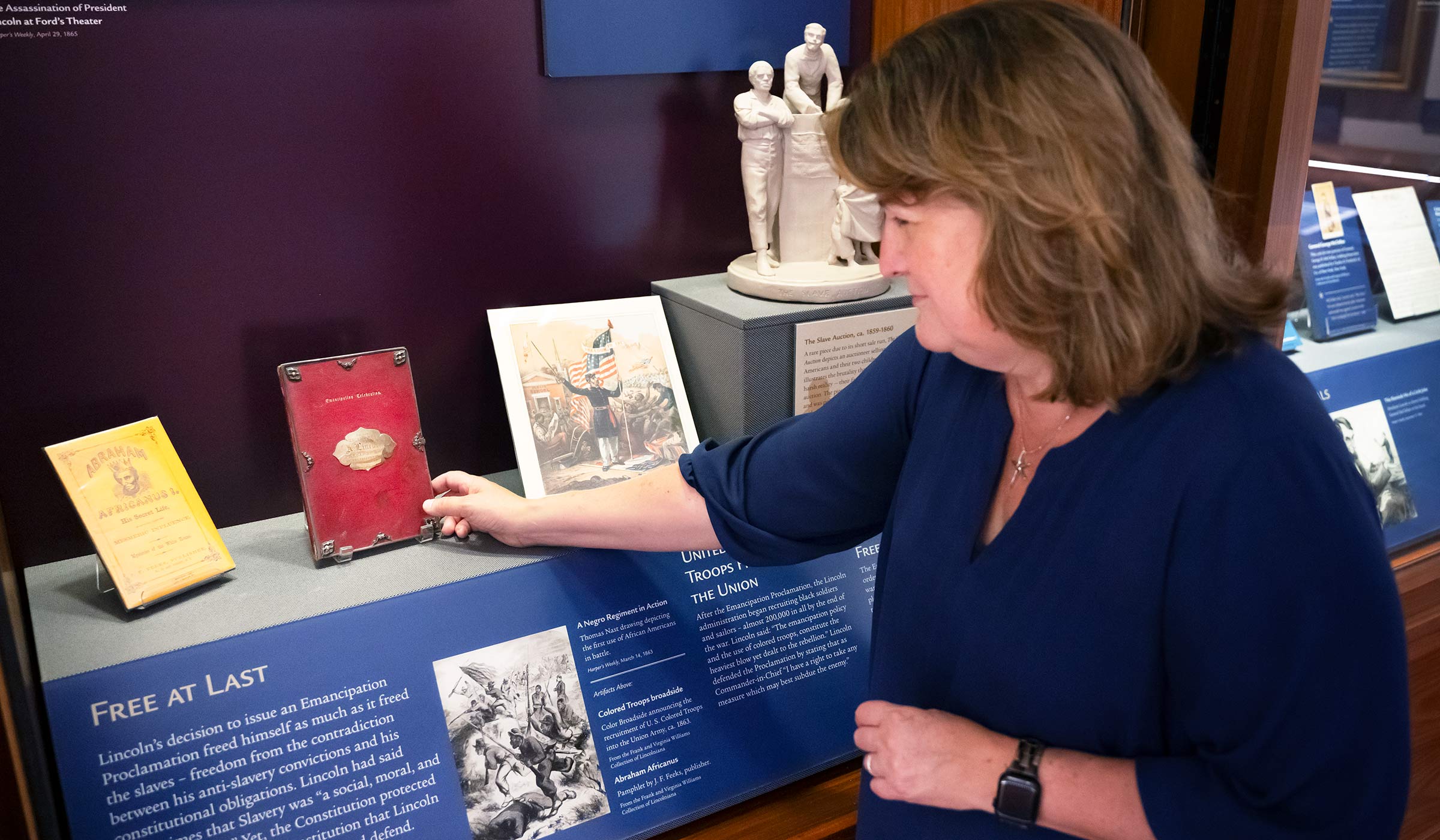 Woman in blue reaching into wood encased display towards red book. 