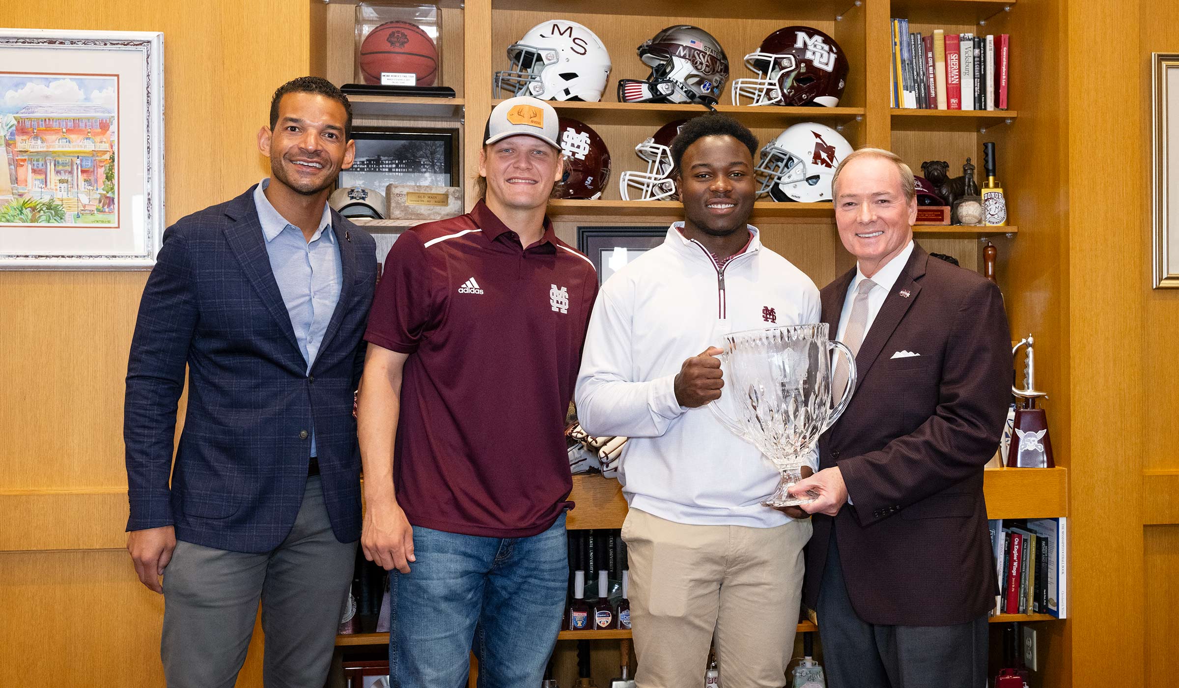 Four men with a glass trophy in front of bookshelf of football helmets