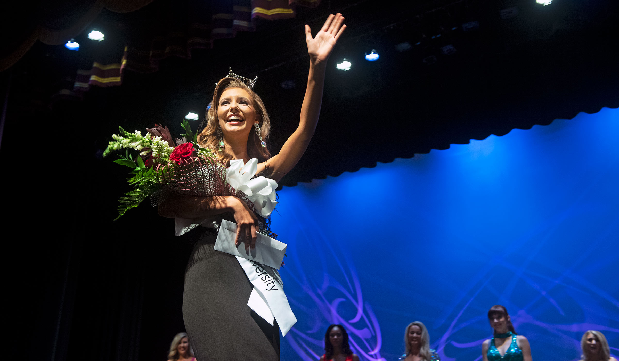 Female student in black evening gown with white sash, roses and crown waving at crowd from stage