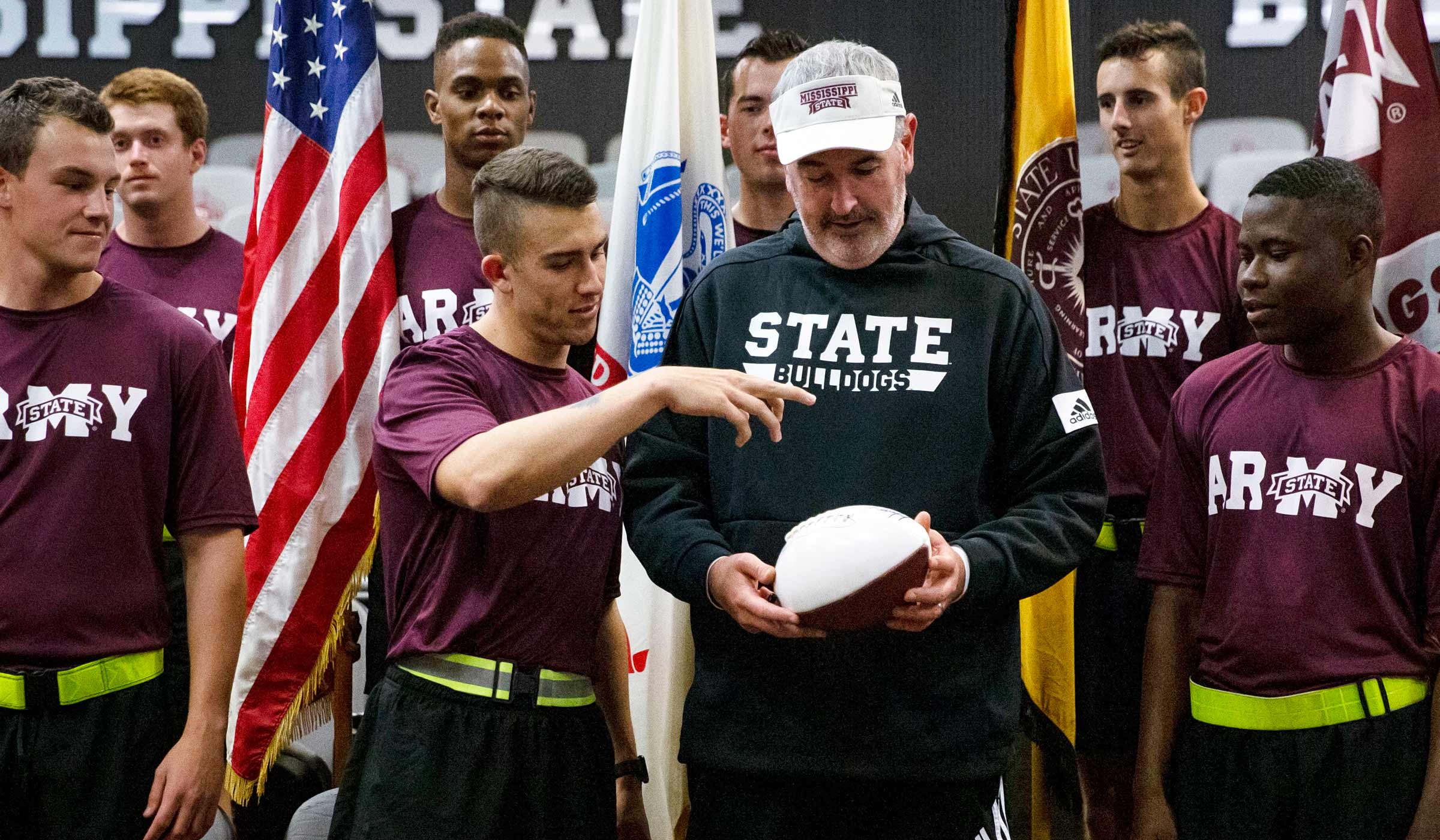 Man in black sweats holding football with Army ROTC in maroon shirts and flags.