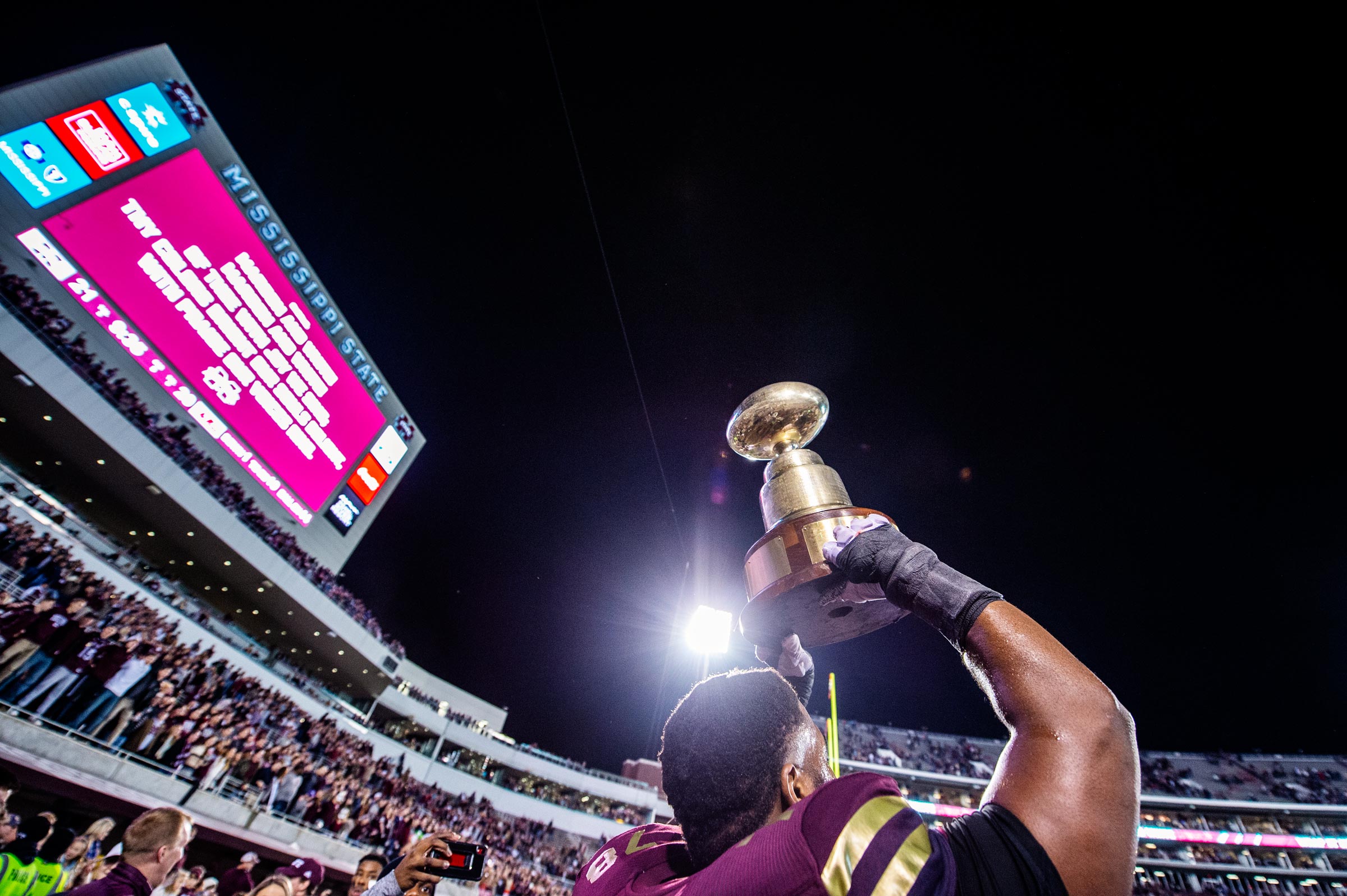 With the nighttime Davis Wade Stadium wrapping around him, an MSU player holds the Egg Bowl trophy high over his head in celebration