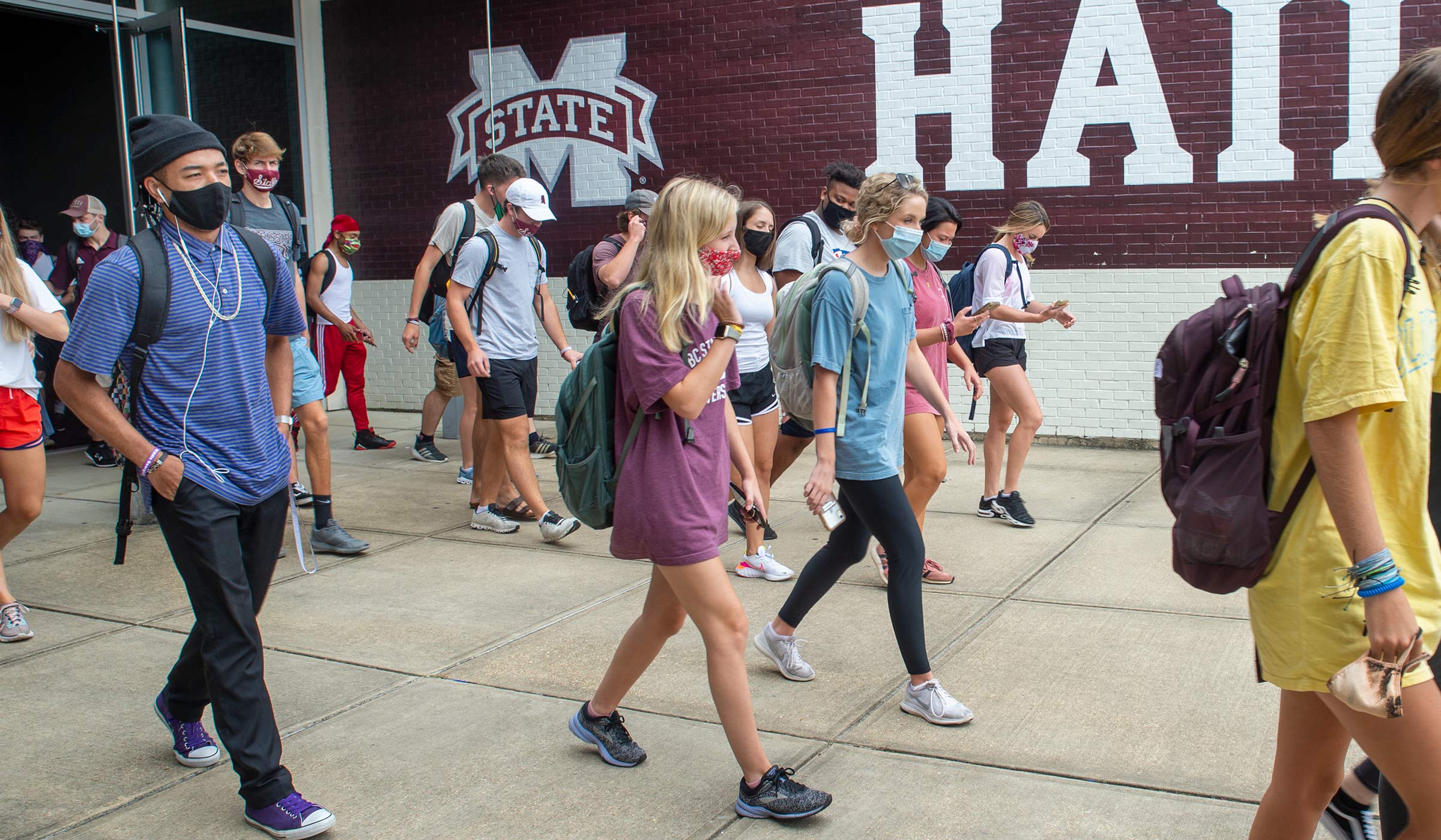With a bit of the Hump&#039;s Hail State sign behind them, masked students exit the doors of the Mize Pavillion entrance after class.