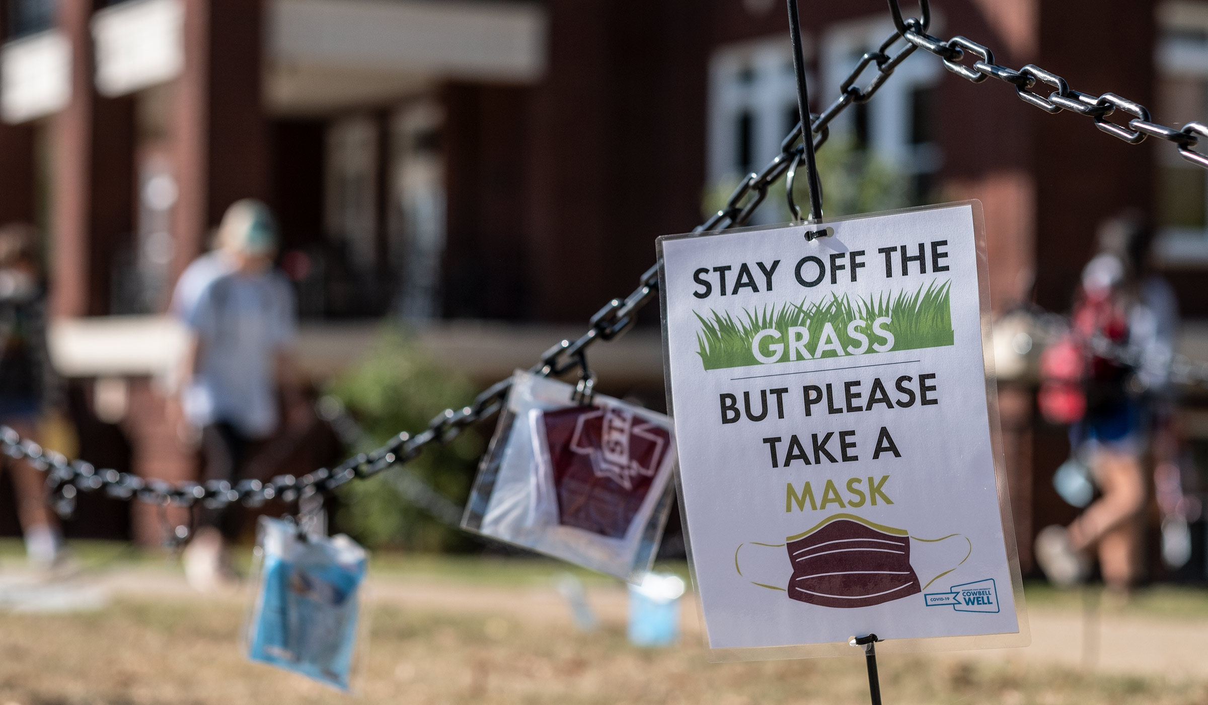Detail of sign hanging on metal chain barrier reads &quot;Stay Off the Grass But Please Take a Mask.&quot;