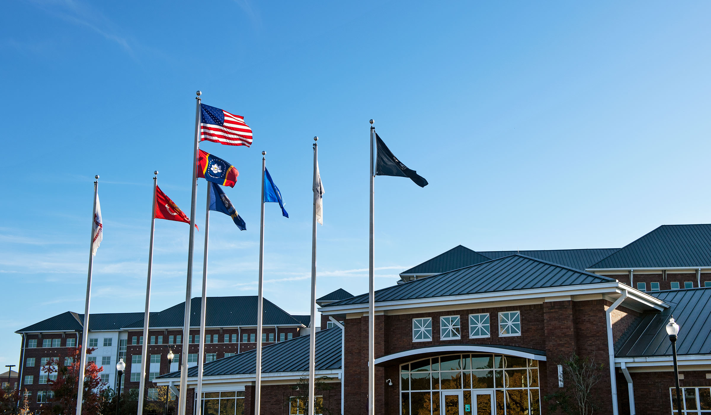 Flags flying in breeze on flagpoles in front of brick buildings. Flags include American flag, new MS state flag, and military flags.