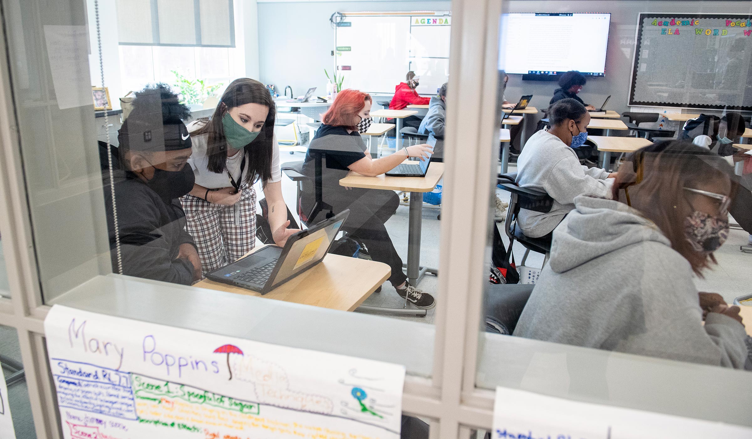 Framed by the windows to the 7th grade classroom, MSU Education major and student teacher Laruen Threadgill bends over a student&#039;s desk to provide to provide guidance.