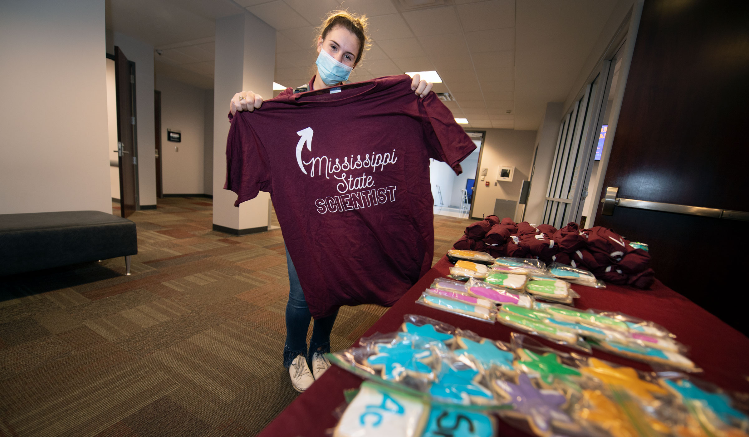 A student holds up a maroon t-shirt that says &quot;Mississippi State Scientist&quot;, with science themed cookies on the table.