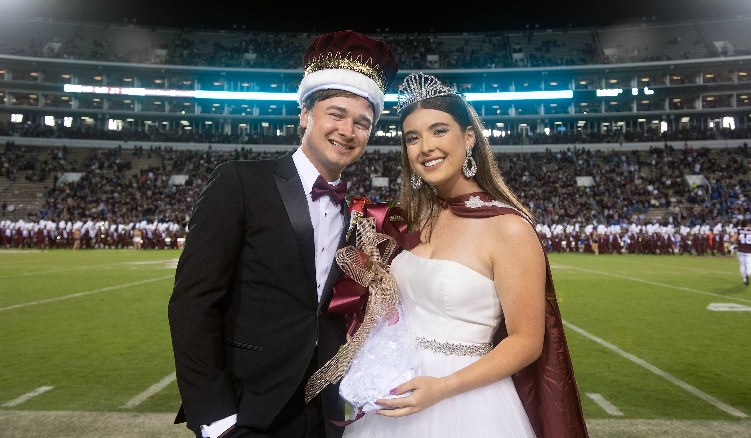 Guy in black tux with maroon bow tie and crown with female in white dress and crown on football field