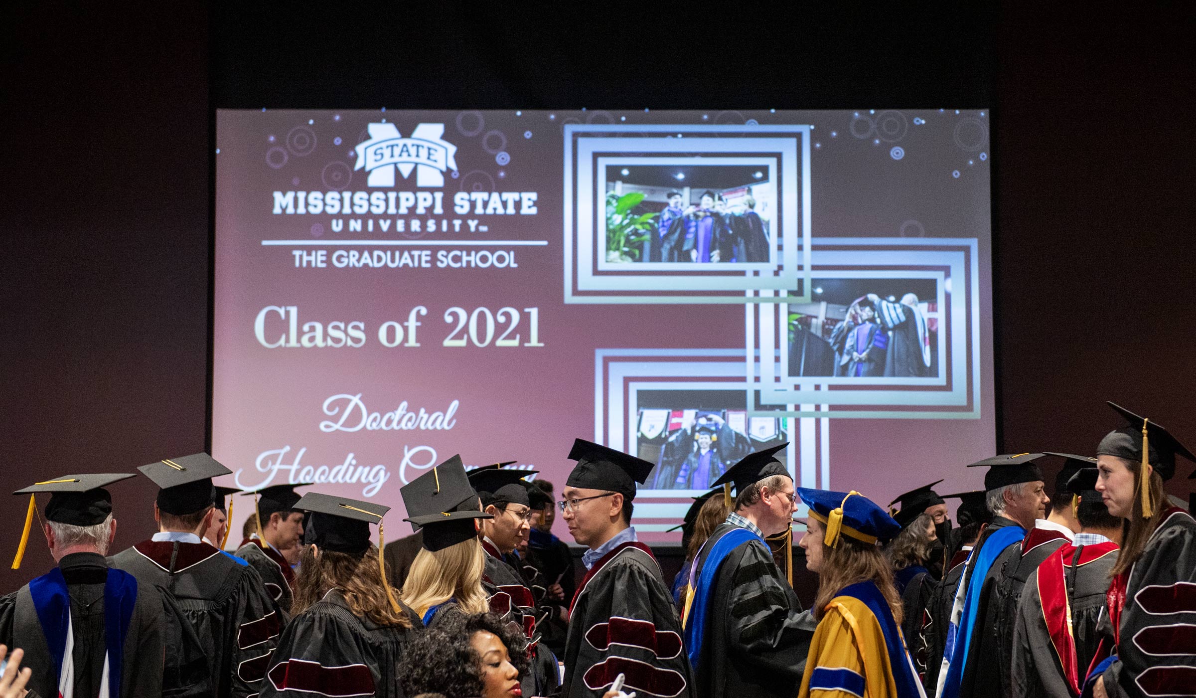 PhD students in full regalia with PhD Hooding Ceremony signage projected on the screen beyond them.