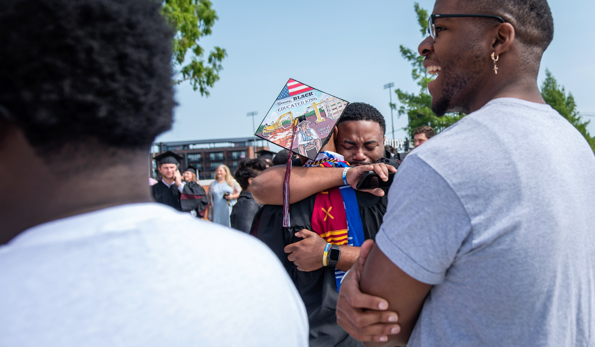 Framed by the shoulders of fellow graduation guests, one student hugs a graduate whose back is turned to the camera, displaying the decorated mortar board he&#039;s wearing.