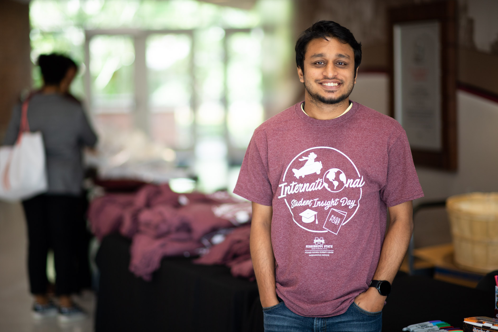 An incoming male South Asian student wearing a light maroon &quot;International Student Insight Day&quot; tee smiles with hands in pockets 
