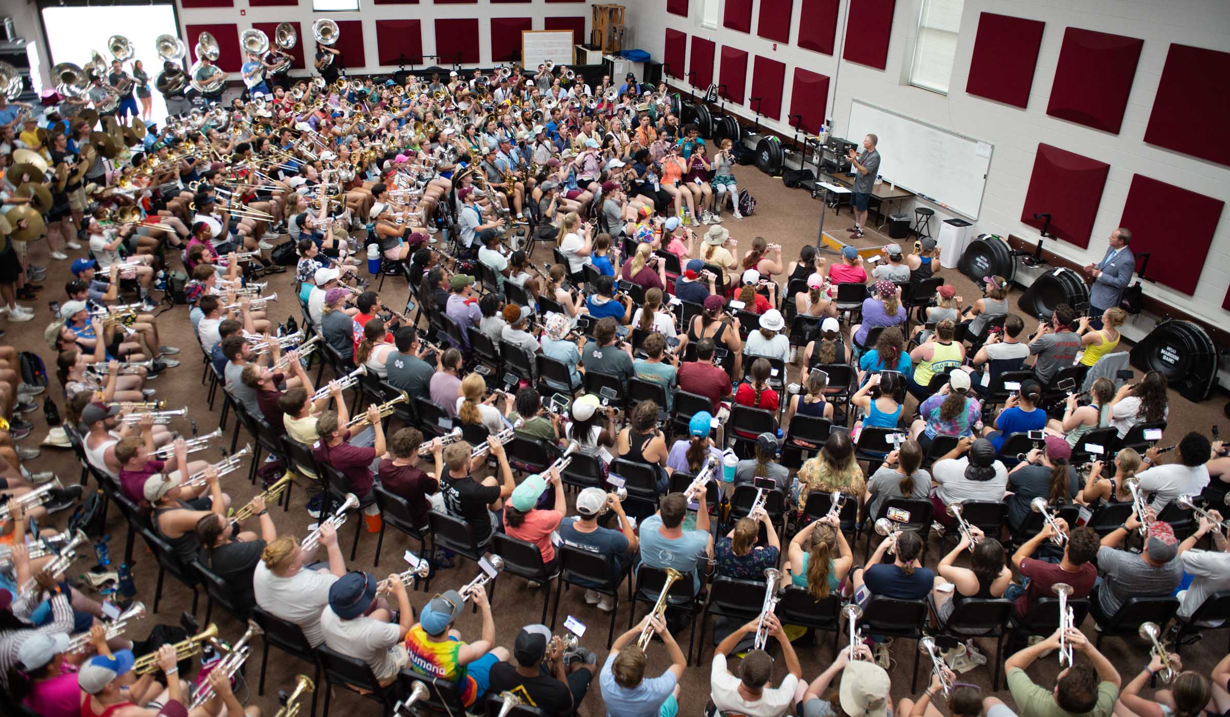 Viewed from above: Band associate director Clifton Taylor conducts a performance of Hail State in the Band Hall, With band members arranged into a semi-circle of seats around him. President Keenum looks on at the far right.