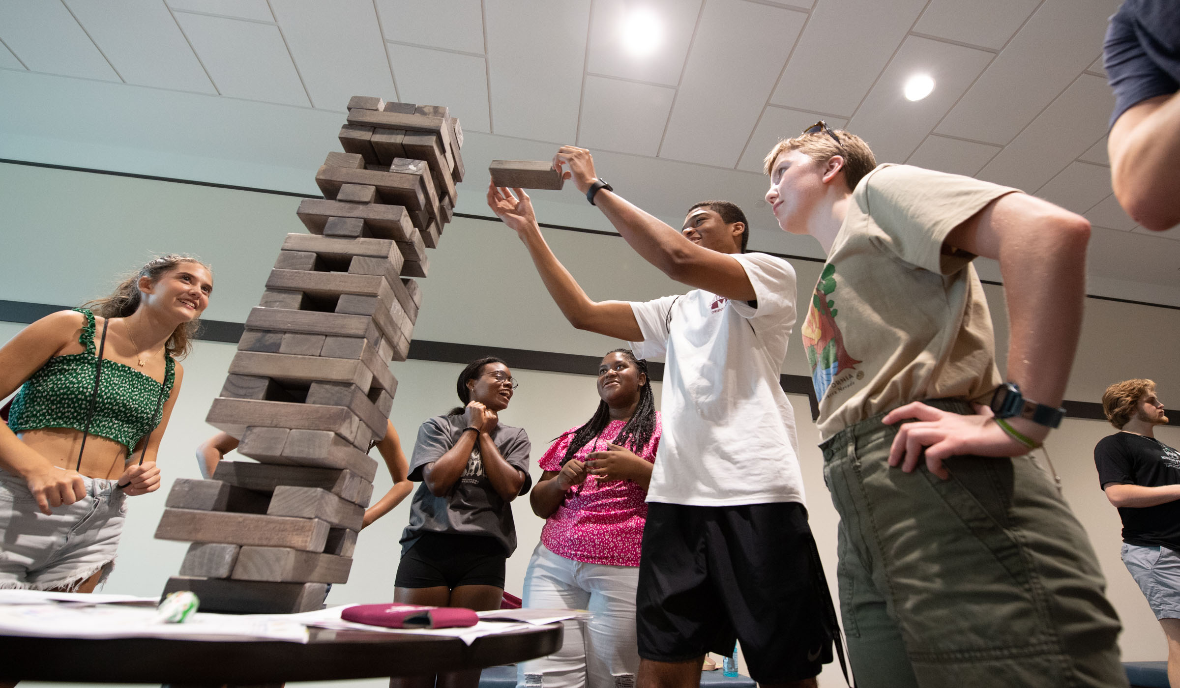 A student reaches to add a block to a tall Jenga tower balanced on a tabletop while other students cheer him on.