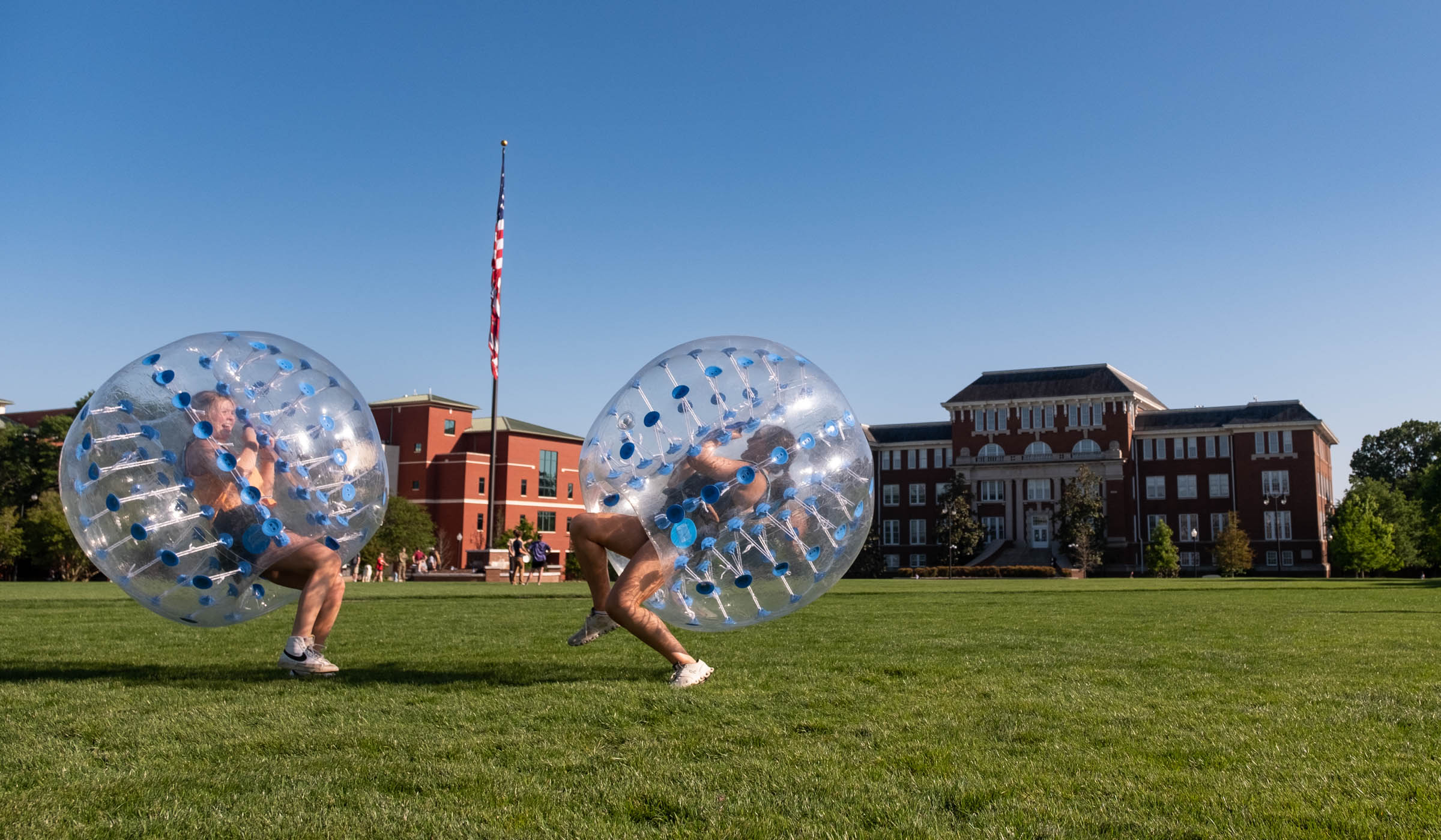 Wearing inflatable bumper bubbles, two students bounce back from eachother on a sunny afternoon on the Drill Field.