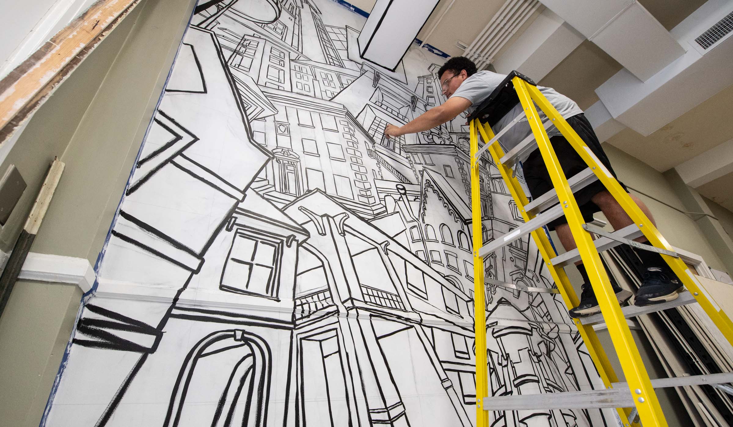 A painted black and white mural covers a wall with a cacophony of MSU residence halls graphically depicted and to the right of the frame, the artist student reaches out to work from the top of a yellow ladder.