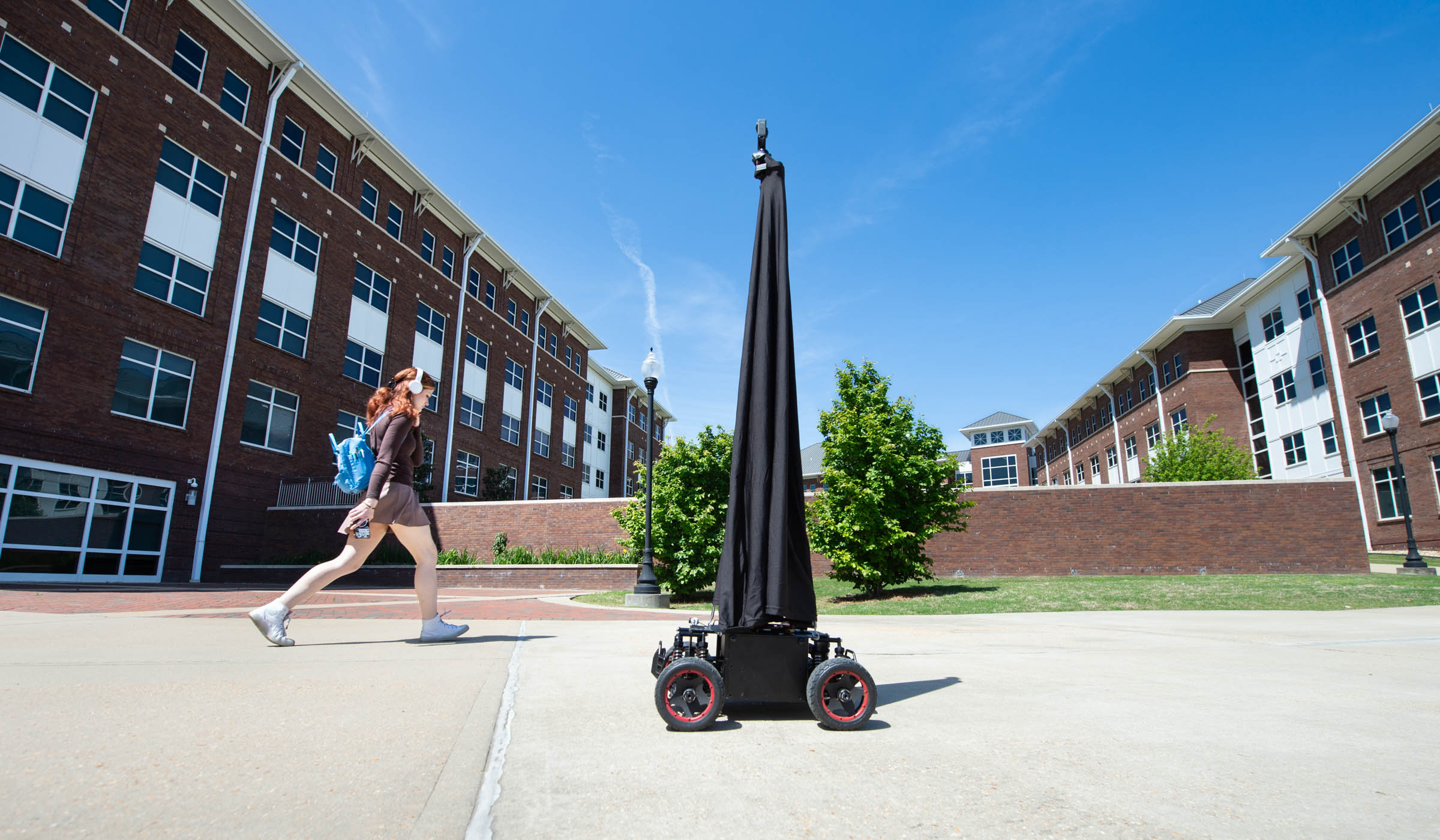 A small black remote-powered vehicle with a black cloth covered pole and camera atop moves down a sidewalk with Dogwood and Deavenport Halls and blue sky behind.