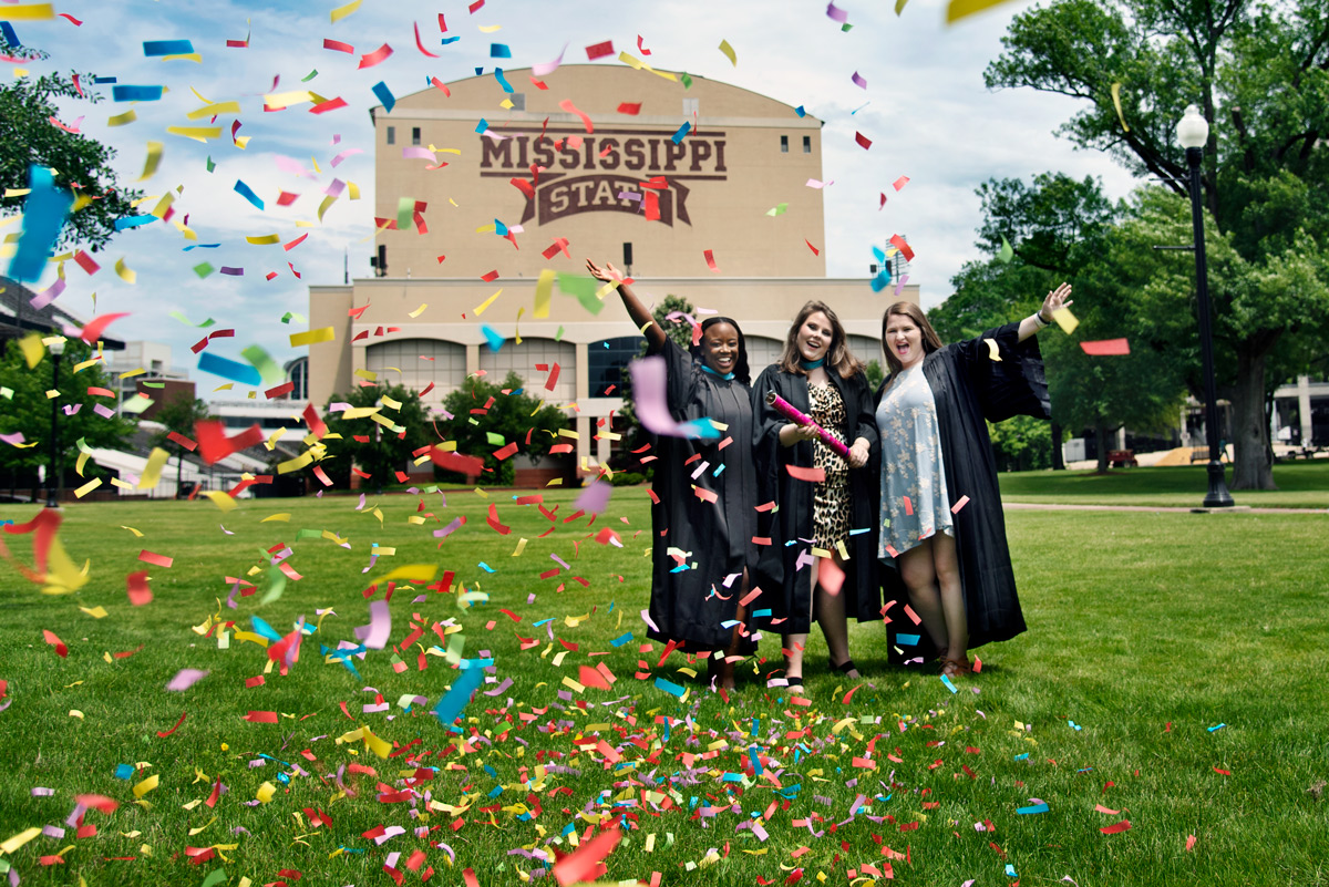 Three graduates pose for photos in the Junction with confetti falling all around them.