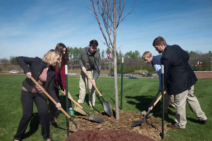 Five students shovel dirt for a freshly planted tree near Highway 182.
