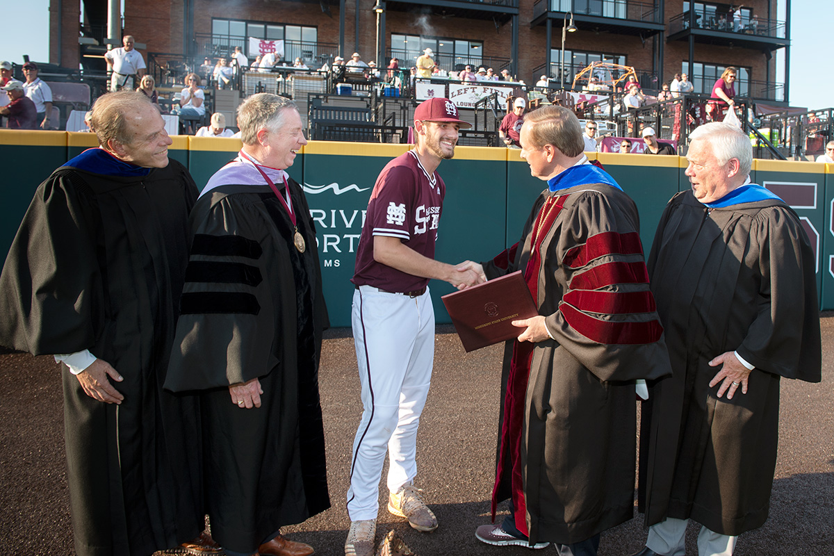 Men in graduation regalia on baseball field shaking hands with baseball player holding diploma