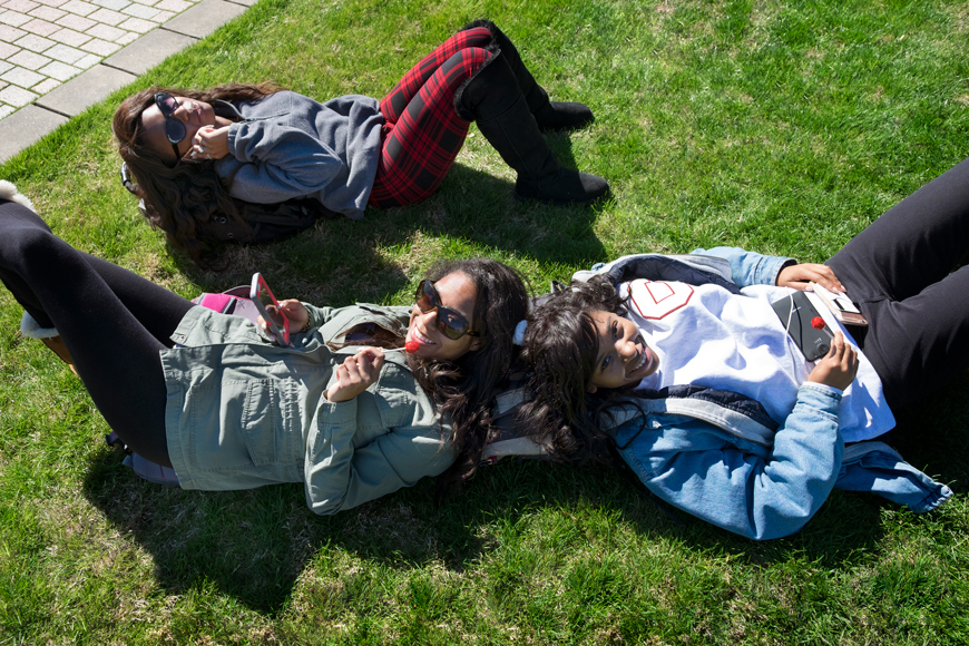 Three friends soak in the sunshine on the Drill Field grass: two with lollipops, one with a cellphone.