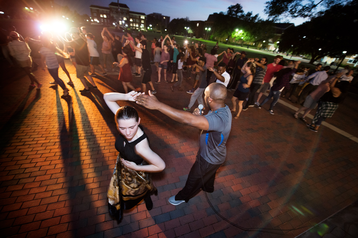 At dusk on the Drill Field, 2 graduate students demonstrate a salsa spin, with dancers practicing in the background.