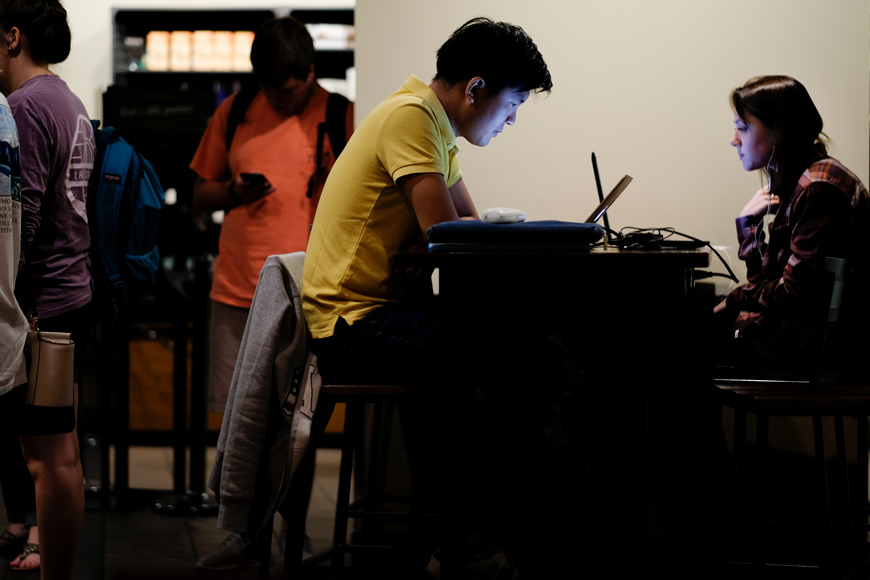 Students Dukjae Lee and Haley Jenkins study in Starbucks, with their faces lit up by their computer screens as they sit on opposite sides of table.