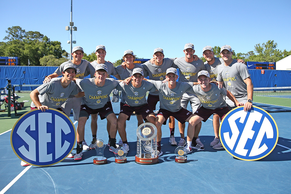 12 men with SEC signs and multiple trophies in front of them posed on blue tennis court