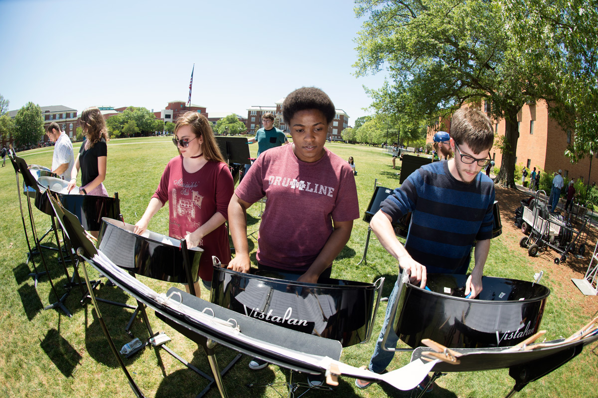 Five students play steel drums in the foreground on a Drill Field sunny day.