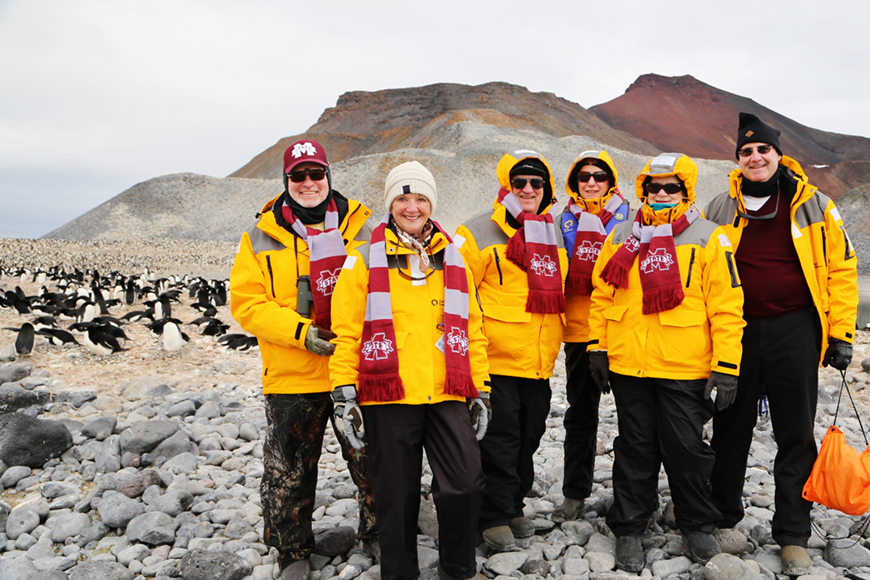 Three couples dressed in yellow jackets with MSU maroon accessories pose near Adele penguins in Antarctica.