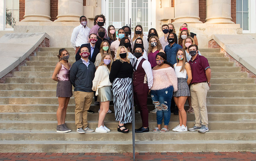 A group of students wearing masks stand on the steps of Lee Hall.