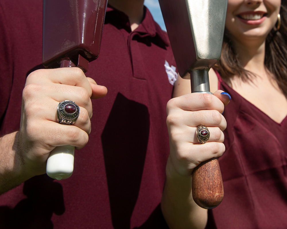 Close up of a man and woman in maroon holding cowbells while showing their MSU class rings
