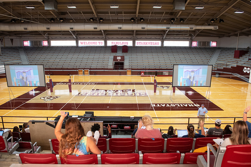 John Blanton teaches an animal and dairy science on a volleyball court as students raise their hands in the stands.