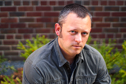 Benjamin Percy stares at the camera while seated in front of a brick wall lined with green plants.