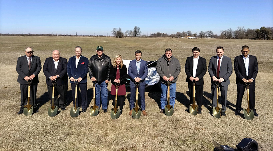A large group stands behind shovels in a large field that will become an industrial facility.
