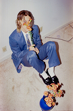 A man in a blue suit sits on carpet near a blue vase of flowers while holding flowers in front of his face.