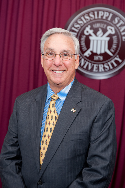 Portrait of Danny Hossley wearing a suit and tie in front of a maroon drape and MSU seal