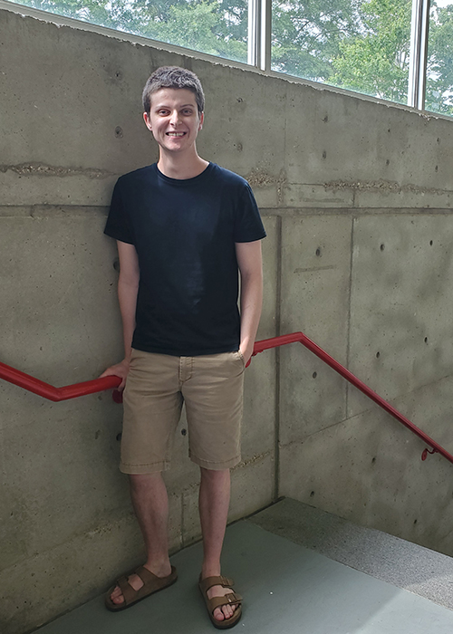 Architecture student John Spraberry smiles while holding a bright red railing in front of a large concrete wall at Giles Hall.