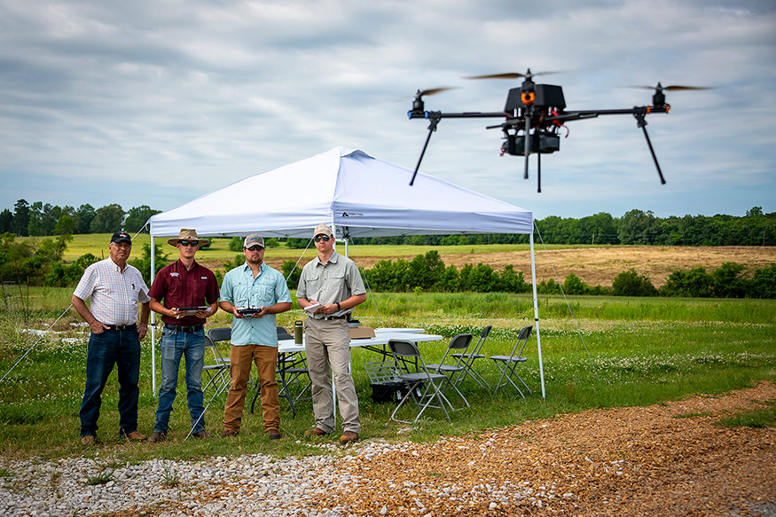 Four men in the background left stand as they control a UAV in the foreground right