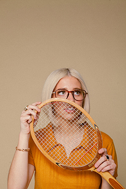 A blonde woman wearing a bright orange shirt and large eyeglasses holds a tennis racket in front of her face.