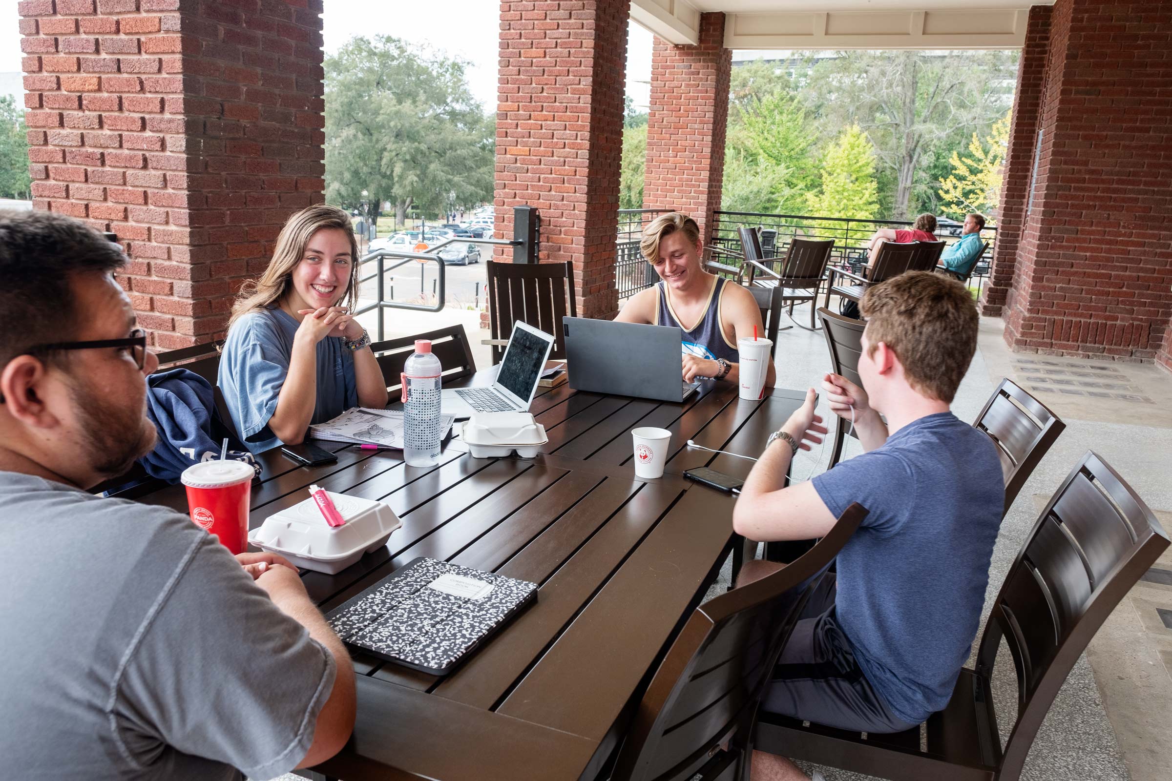 Four students chat and eat lunch between classes at a table on the YMCA&#039;s porch.