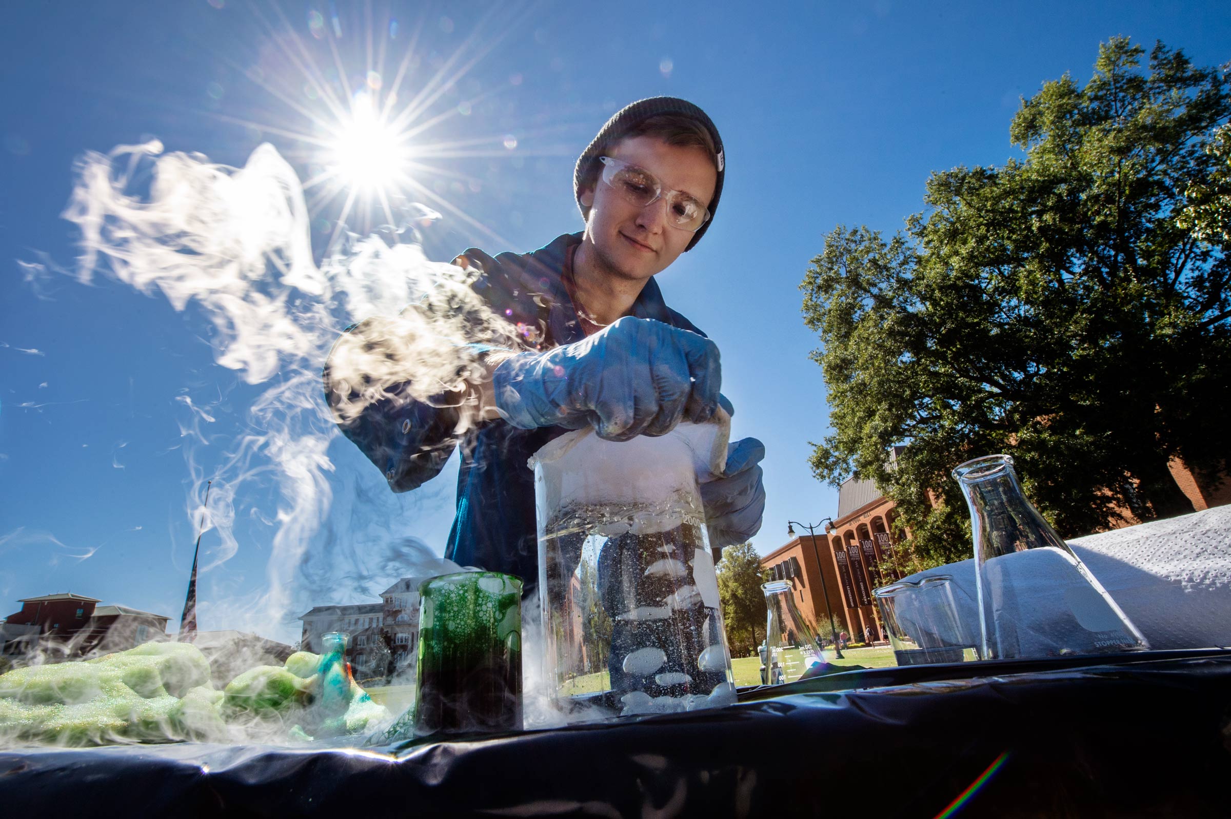 Student Hayden Davis works with beakers of liquid bubbling on Drill Field demonstration table, with sunflare behind.