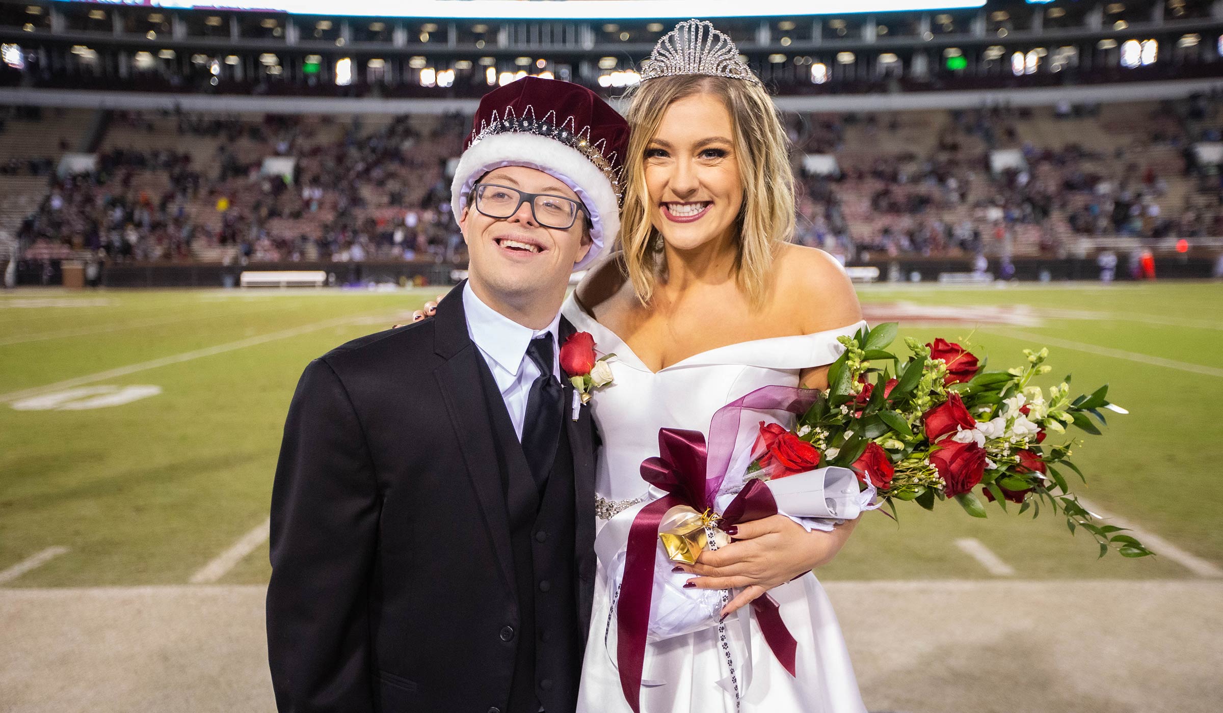 Male in black suit and maroon crown with female in white gown and tiara on sideline of football field.