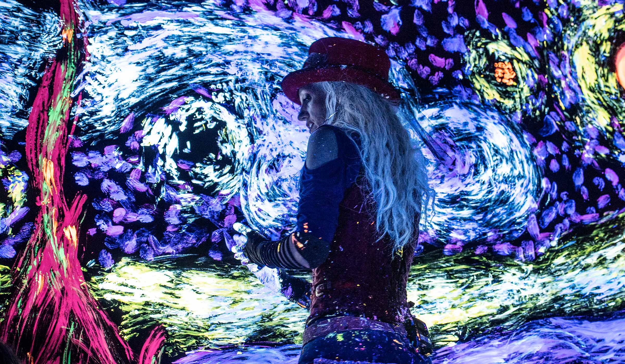 Lit by glowing blaclights, the female Artrageous lead performer uses her hands to add glowing paint to a canvas covered with a a swirly sky painting.