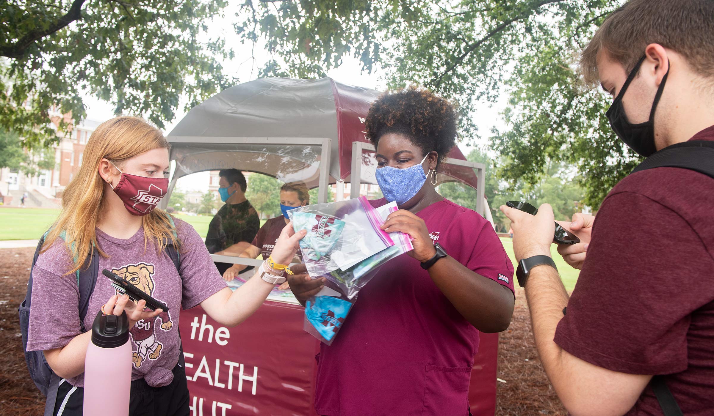 Woman in maroon scrubs handing out masks to two masked students in front of kiosk