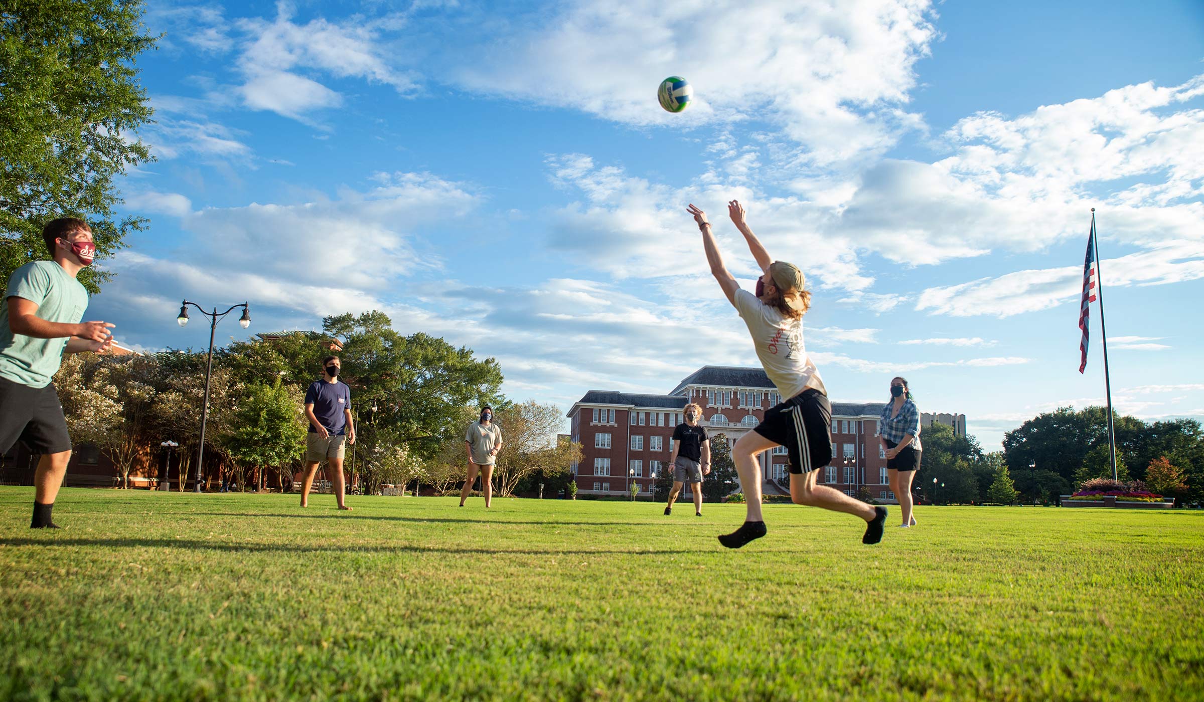 Under a partly cloudy blue evening sky, mask-wearing students toss a volleyball between them on the Drill Field.