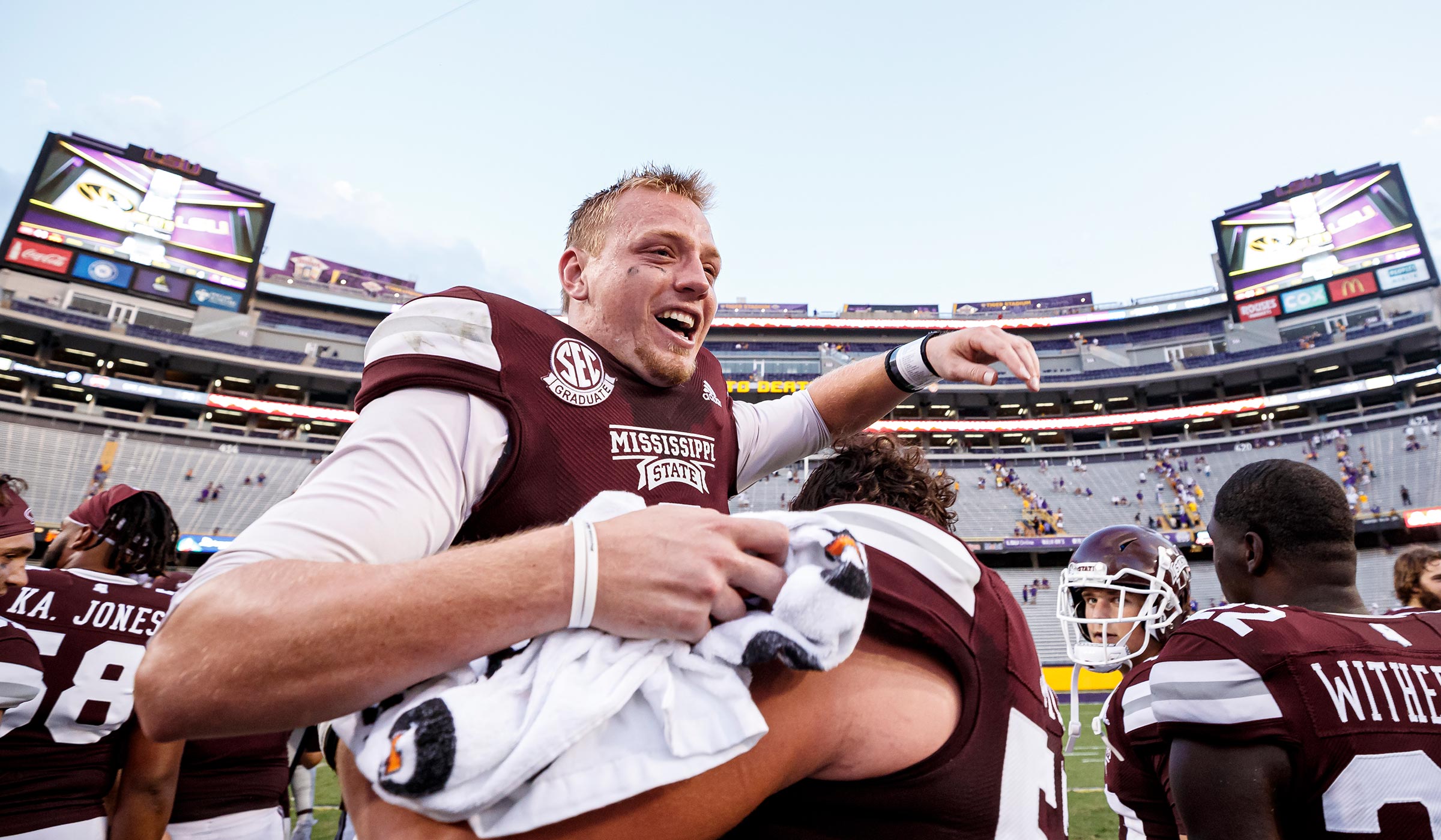 Football player in maroon jersey lifting another player in maroon jersey off ground in celebration 