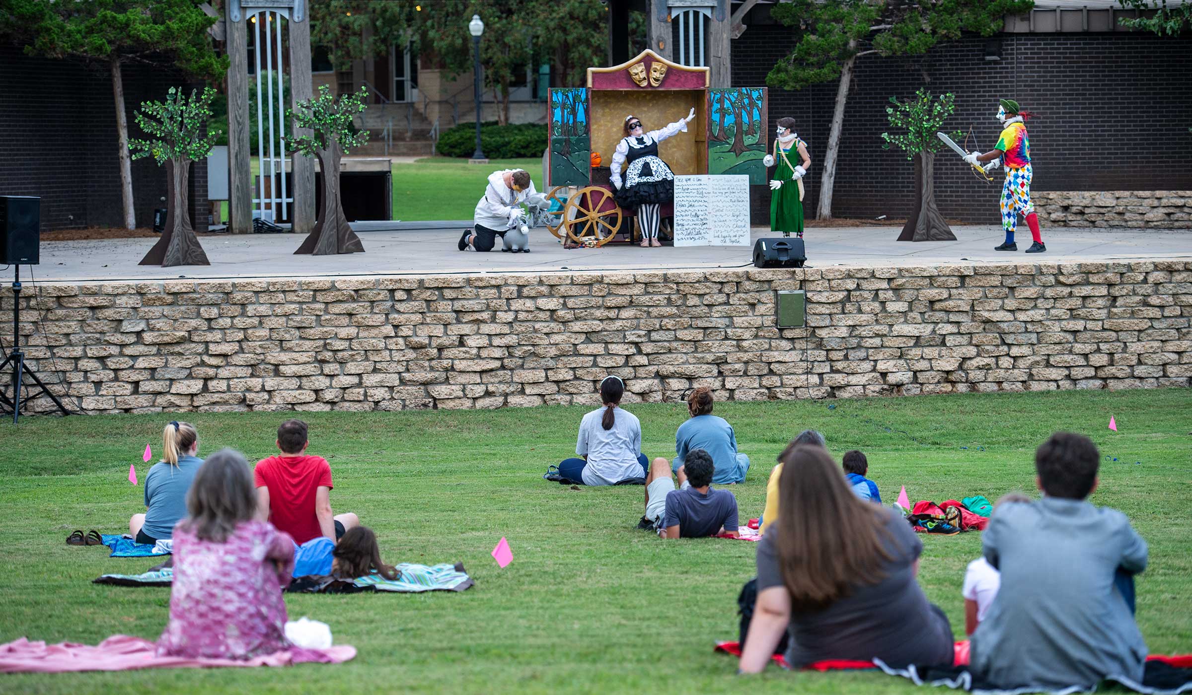 With an audience spread out upon the grass of the outdoor Amphitheater in the foreground, four MSU Theatre actors play the masked characters of Commedia Robin Hood.