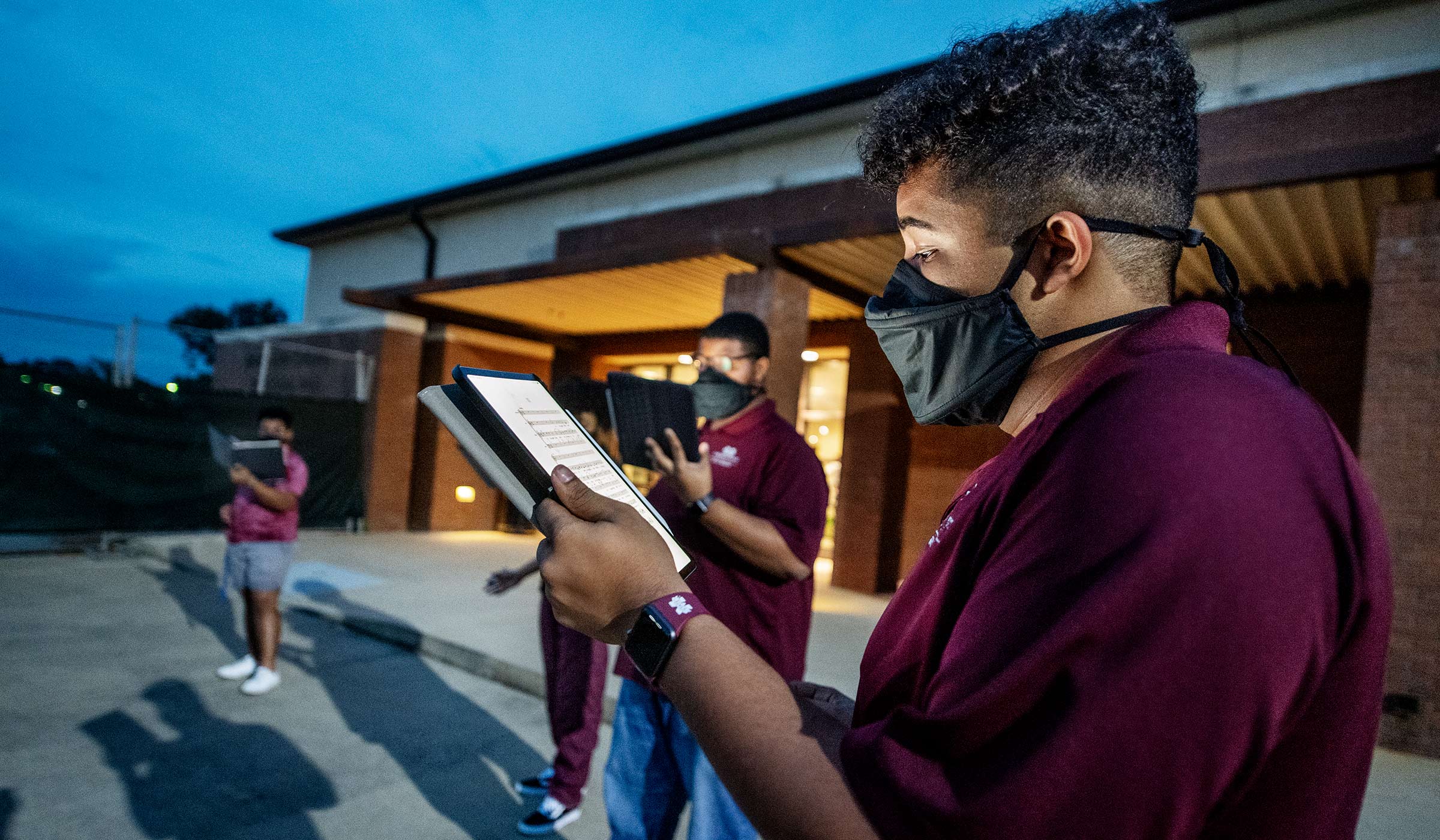 Illuminated by the light of his iPad music, student Ty Lofton and fellow members of the Mississippi State Singers, rehearse outside the Band Hall at dusk, wearing Resonance Masks.