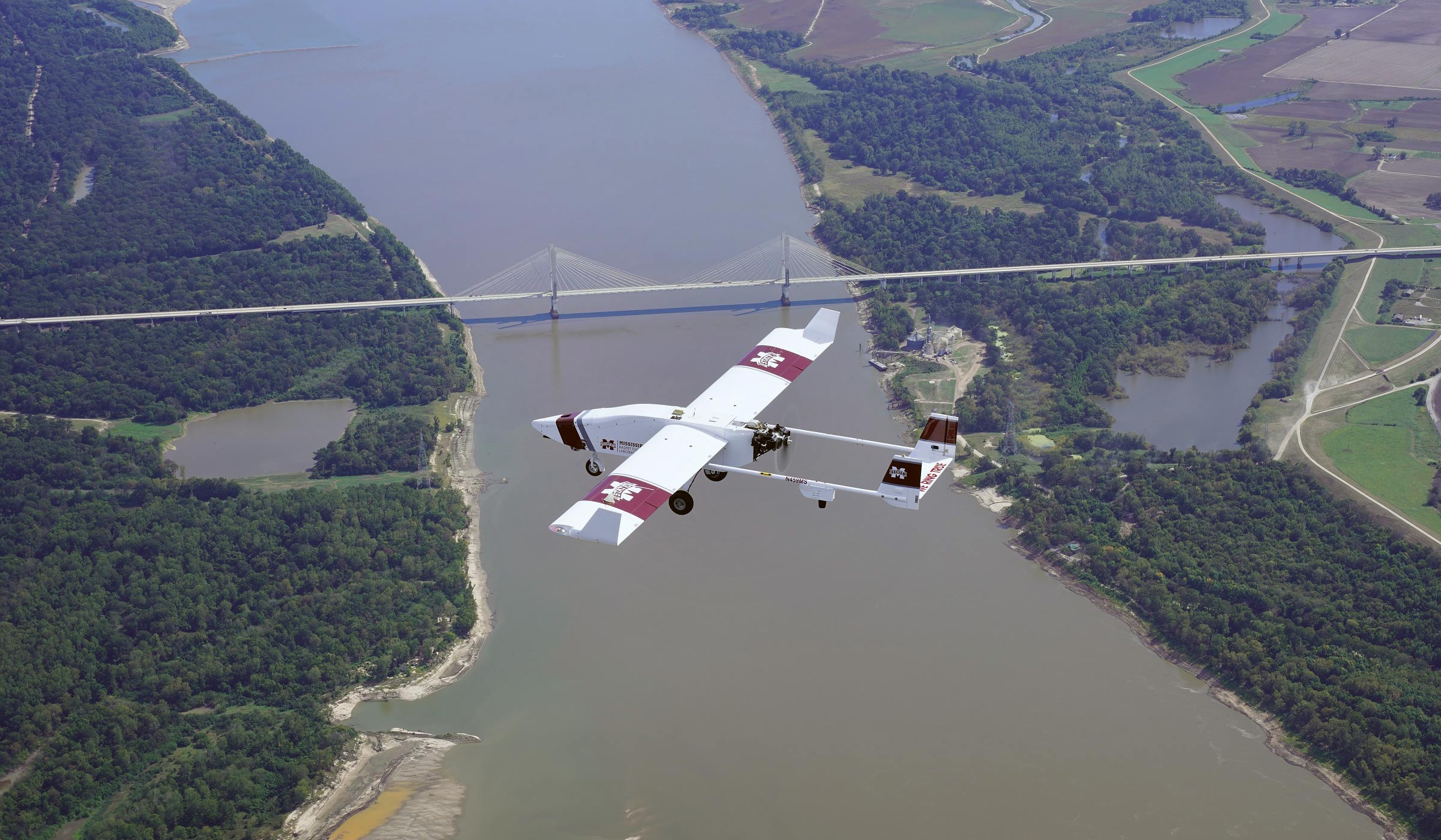 Aerial photo of Rapset UAS Tiger Shark airplane flying with river and bridge below.
