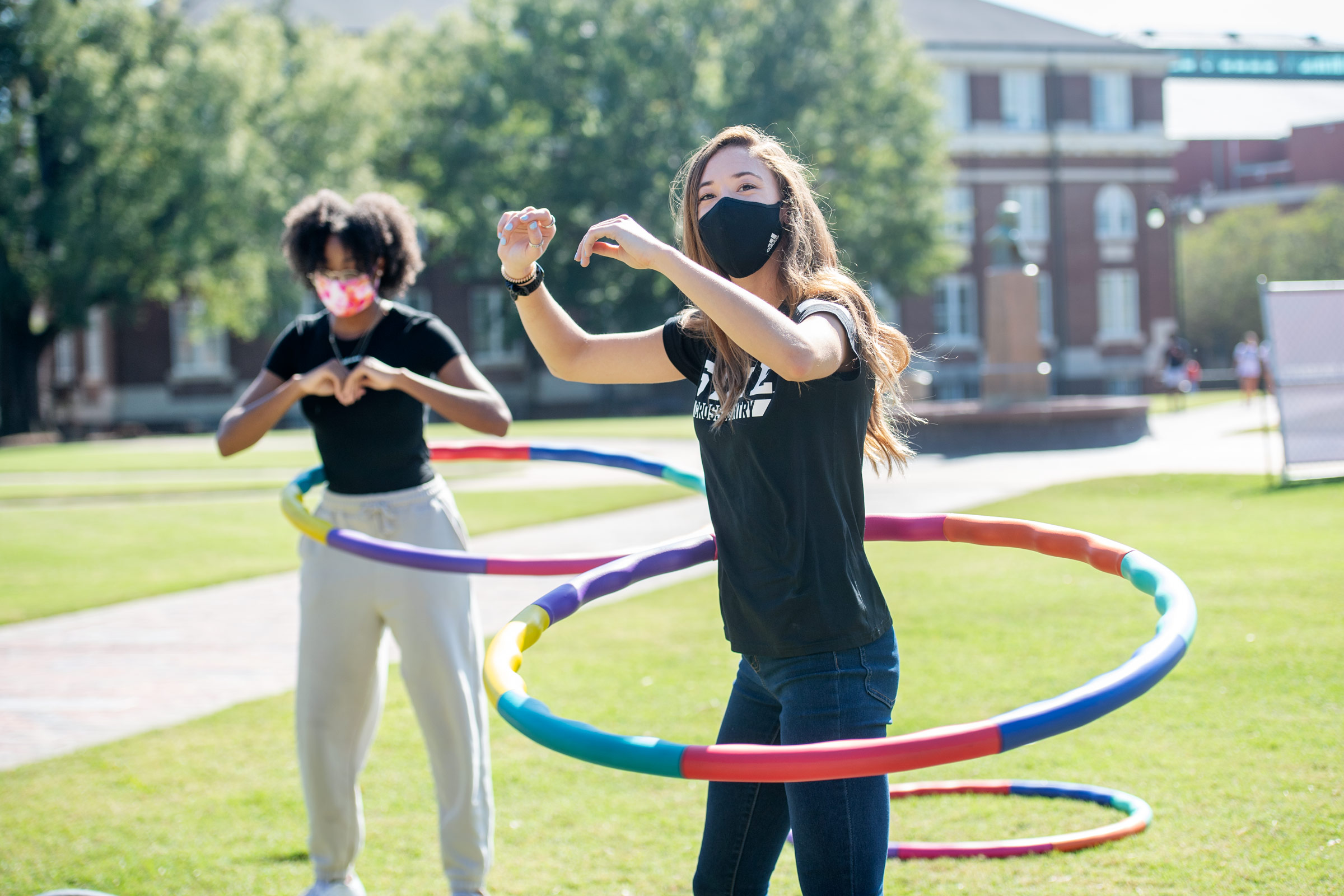 Two masked students hula hoop in the Drill Field sunshine as part of the Pink Dawg Walk event for breast cancer awareness.