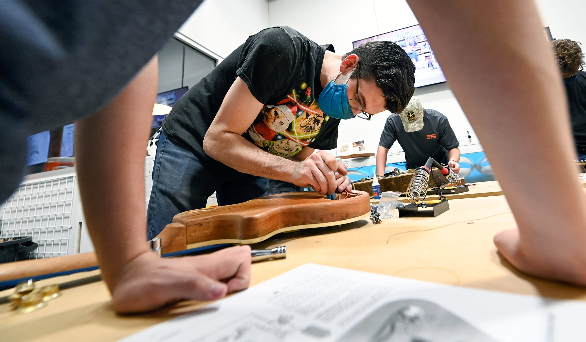 MSU alum and Idea Shop employee Landon Casey works on a guitar, framed by the arms of a participant leaning on the workshop table.